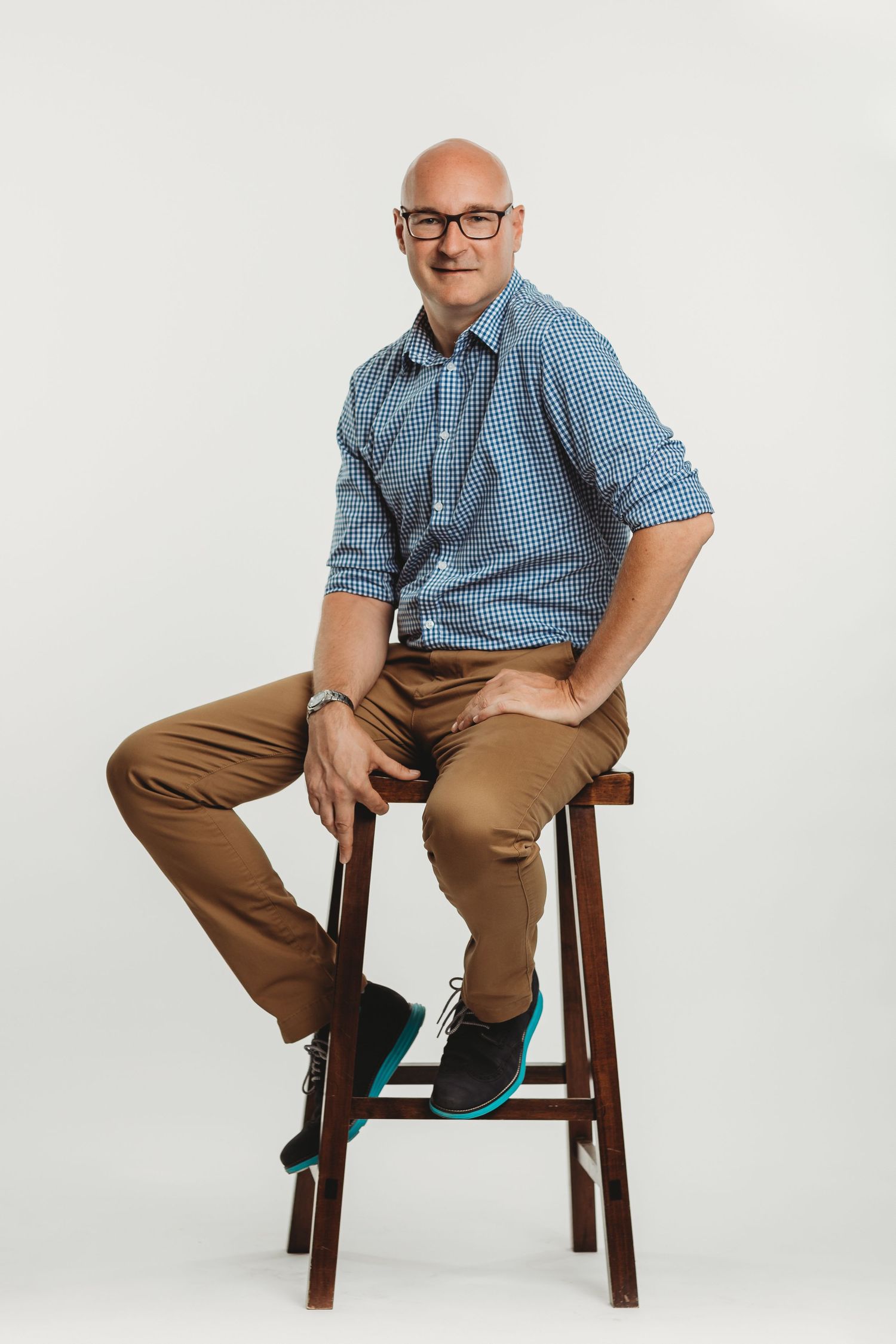 A casual portrait of someone in glasses and checkered shirt sitting on a wooden stool.