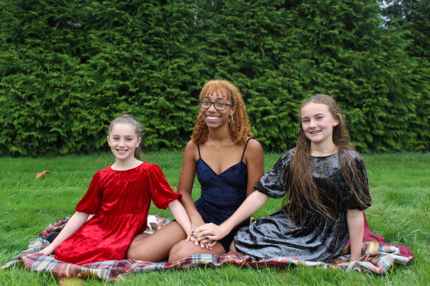 Three friends in elegant dresses posing on a plaid blanket on the grass.