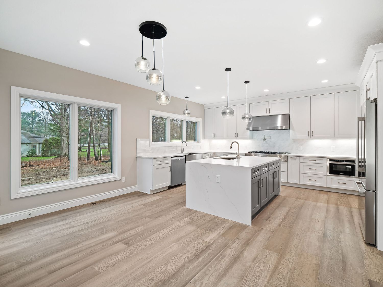 Modern white kitchen with center island, pendant lights, and large windows facing wooded view.