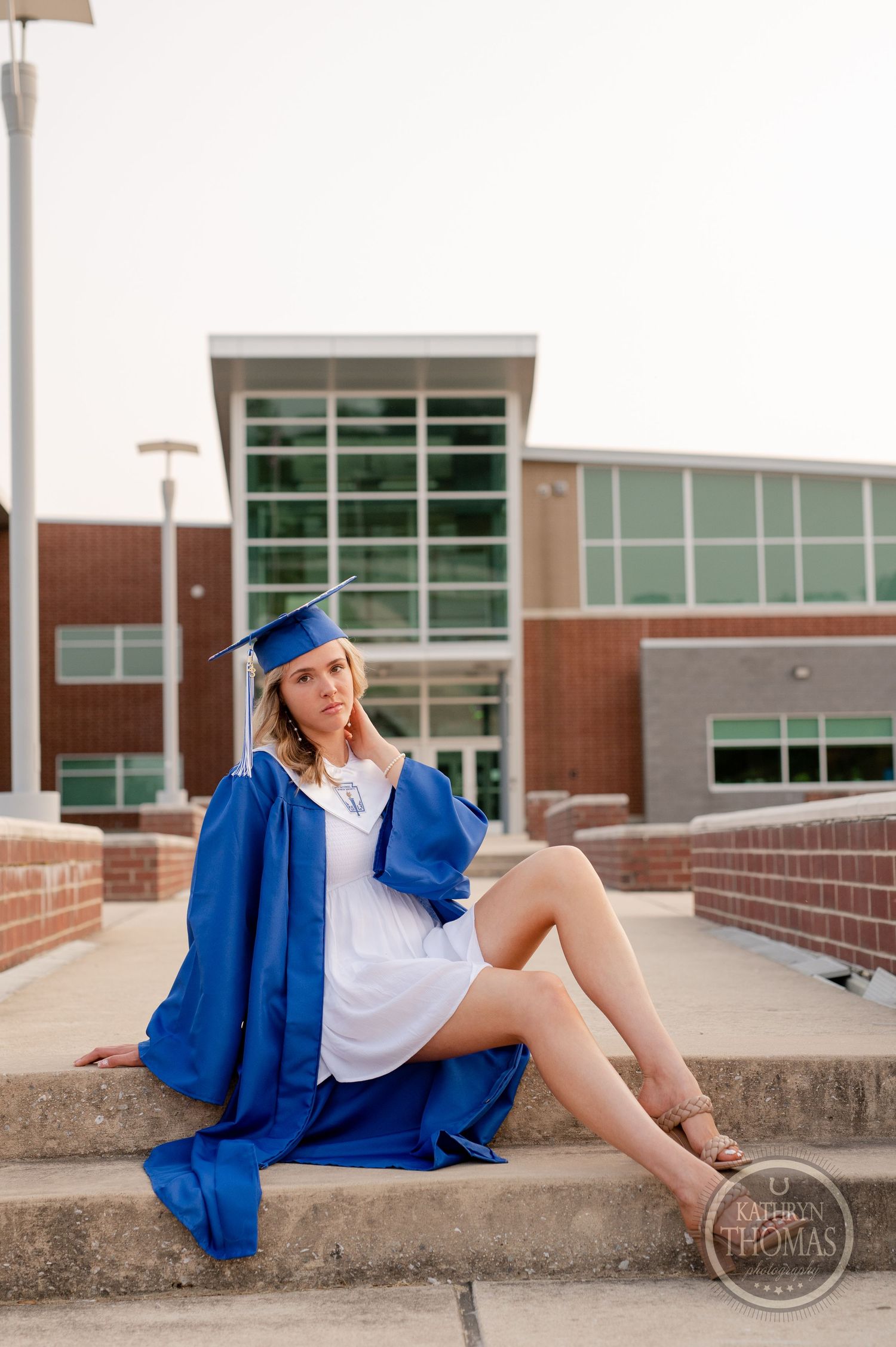 Graduate in blue regalia sits casually on concrete steps with modern building in background.
