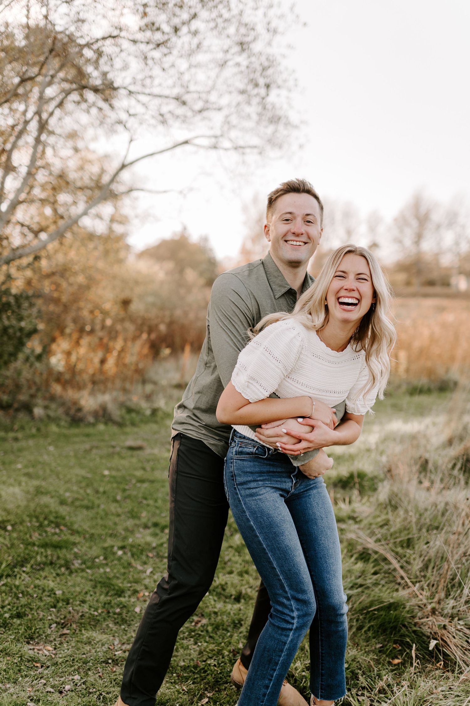 A couple shares a playful moment outdoors in autumn, laughing together against a backdrop of fall foliage.
