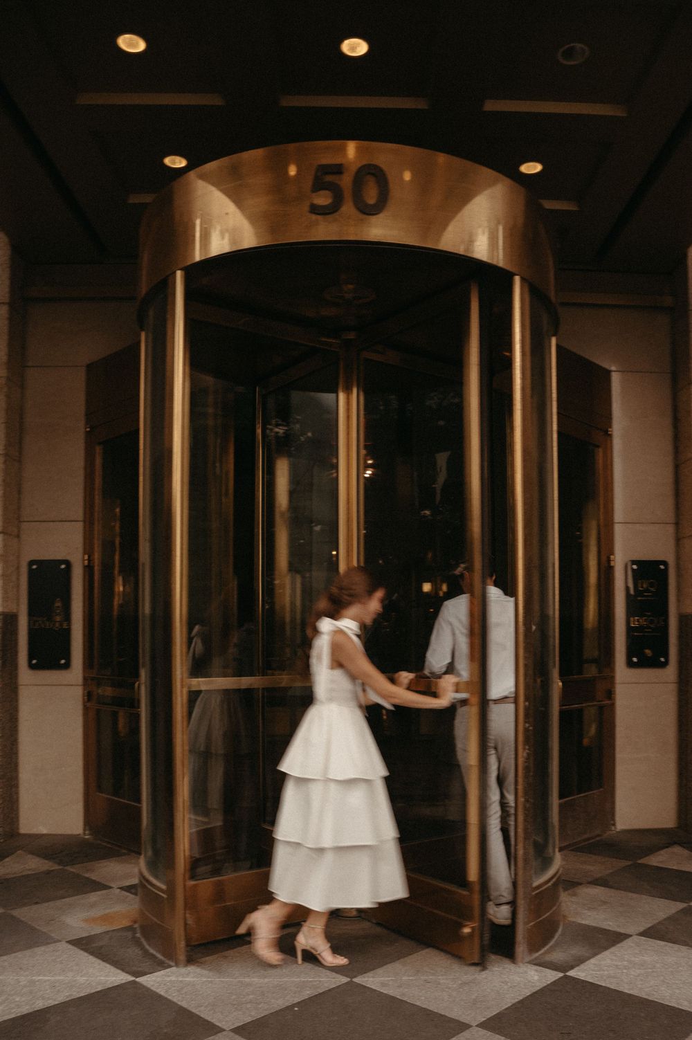 A couple entering a revolving door marked '50' with the bride wearing a tiered white dress.