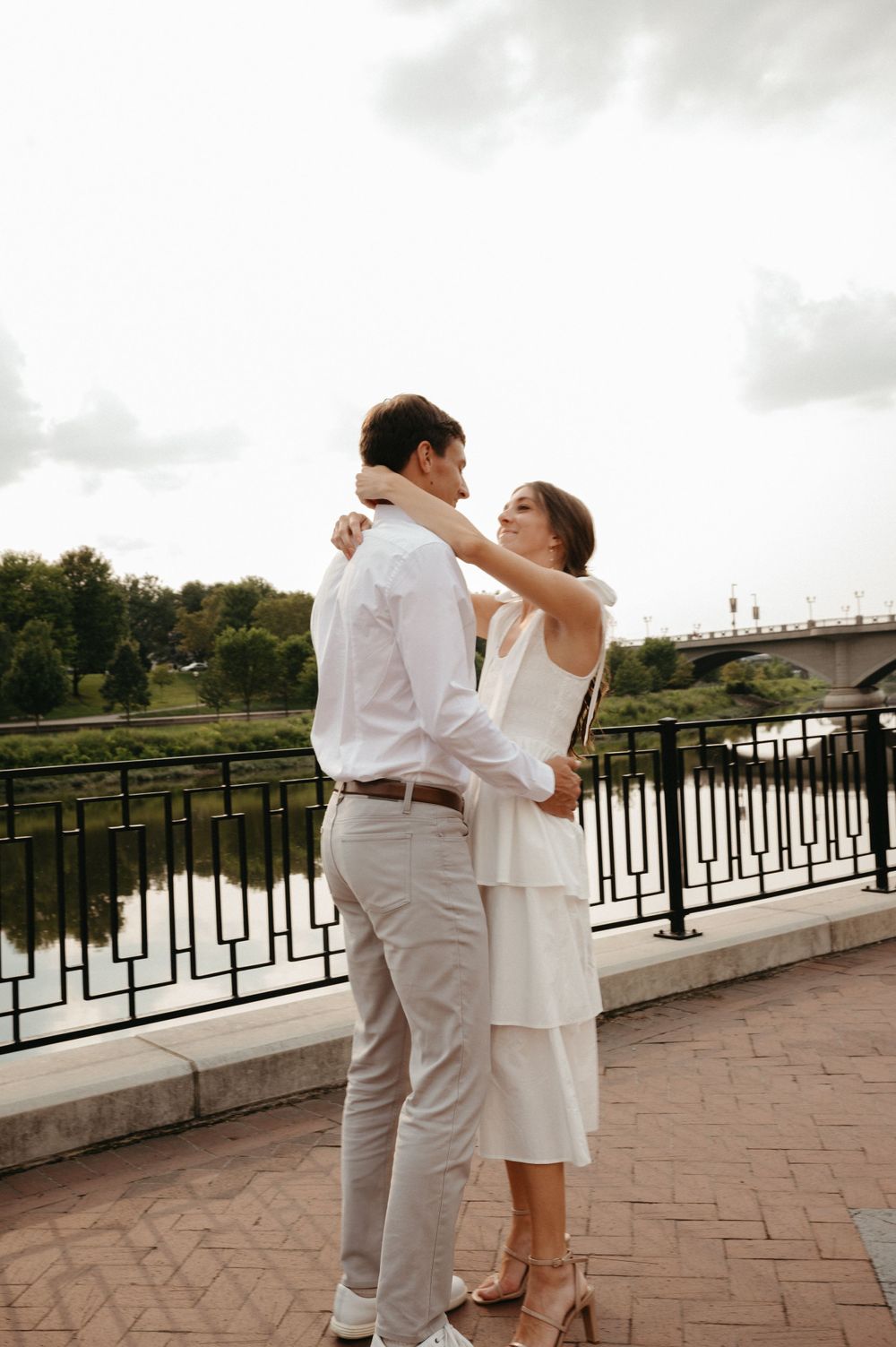 A couple in coordinated white outfits embraces while standing on a brick walkway overlooking a river.
