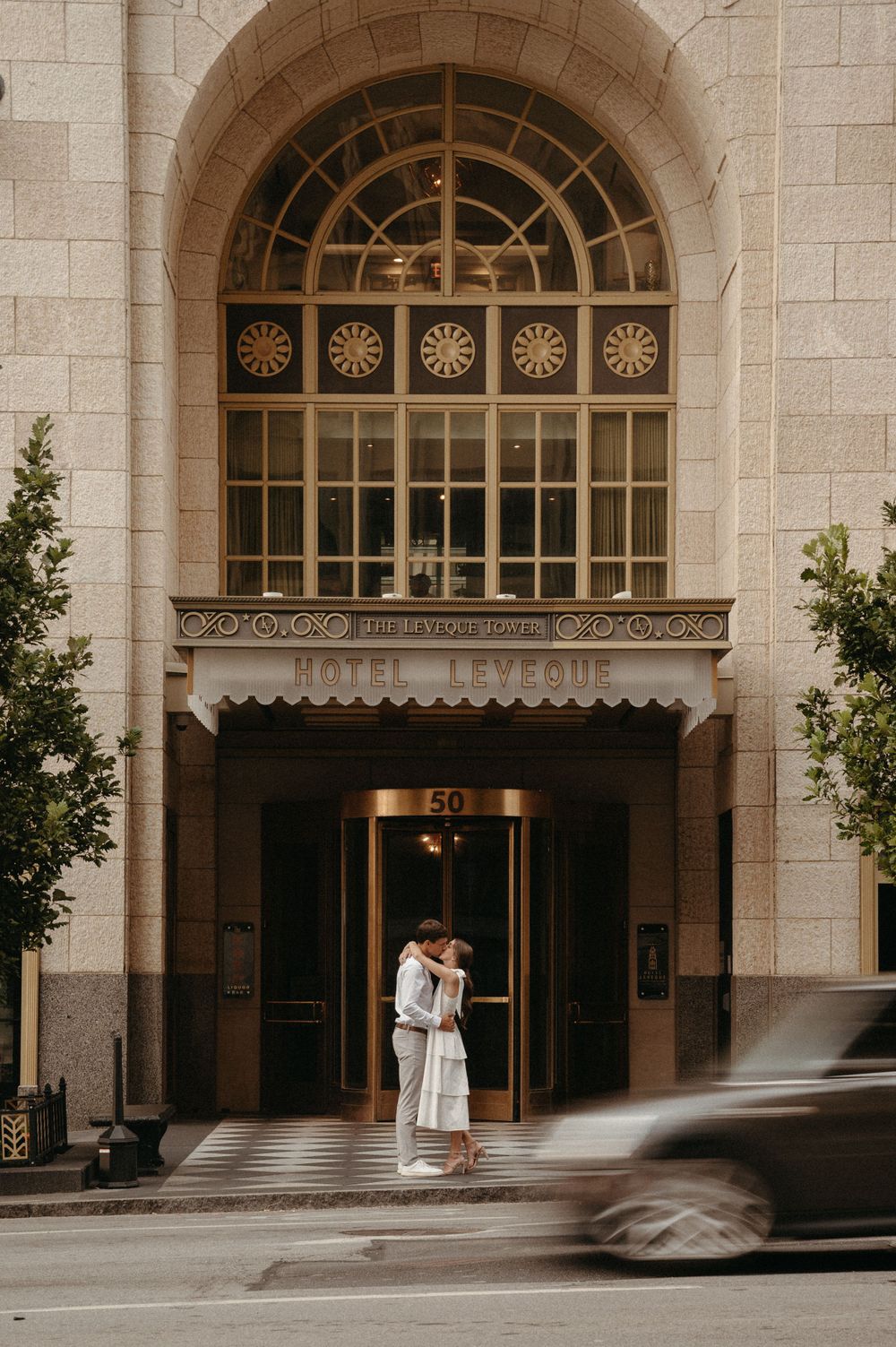 A couple embracing at the entrance of a historic building with an ornate arched window above.