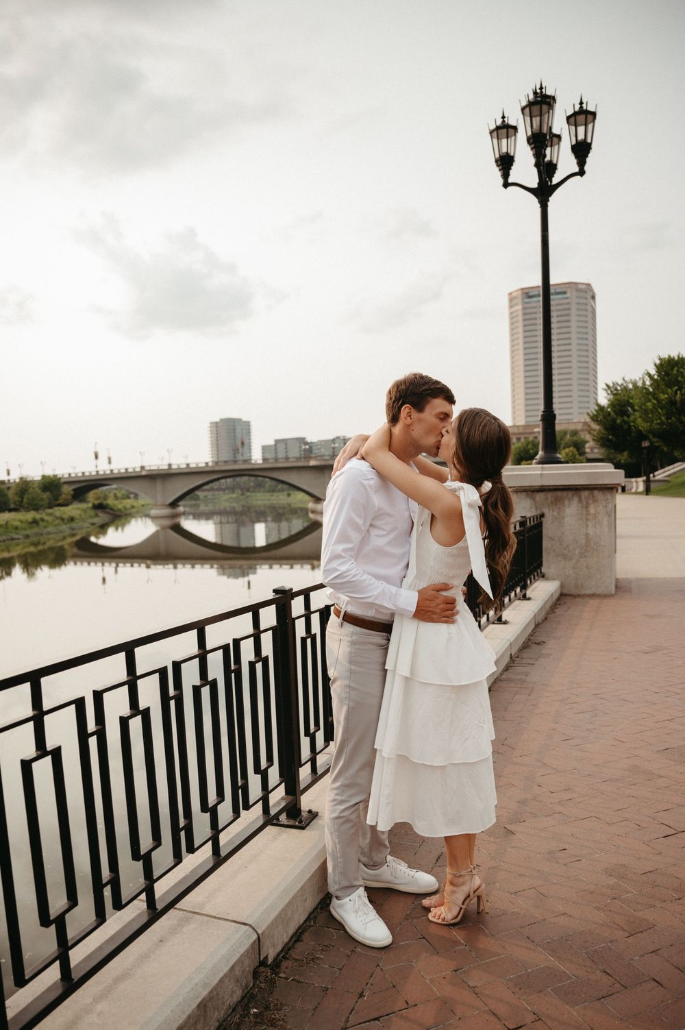 A romantic embrace by the river with an ornate lamp post and bridge in the background.