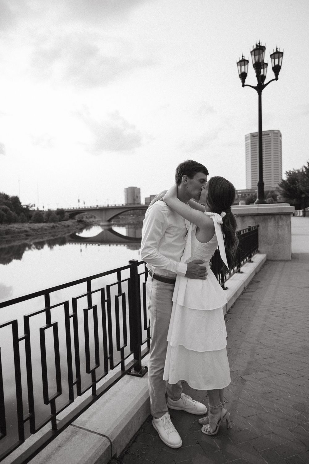 A romantic couple shares an intimate moment on a riverside promenade with vintage lampposts and city buildings in view.