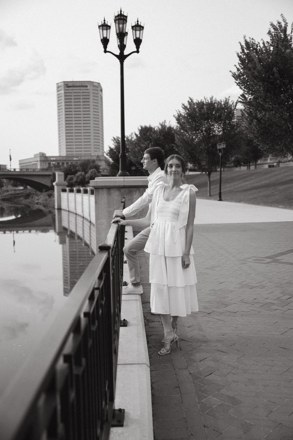 A couple poses along a riverside walkway with an urban skyline in the background.