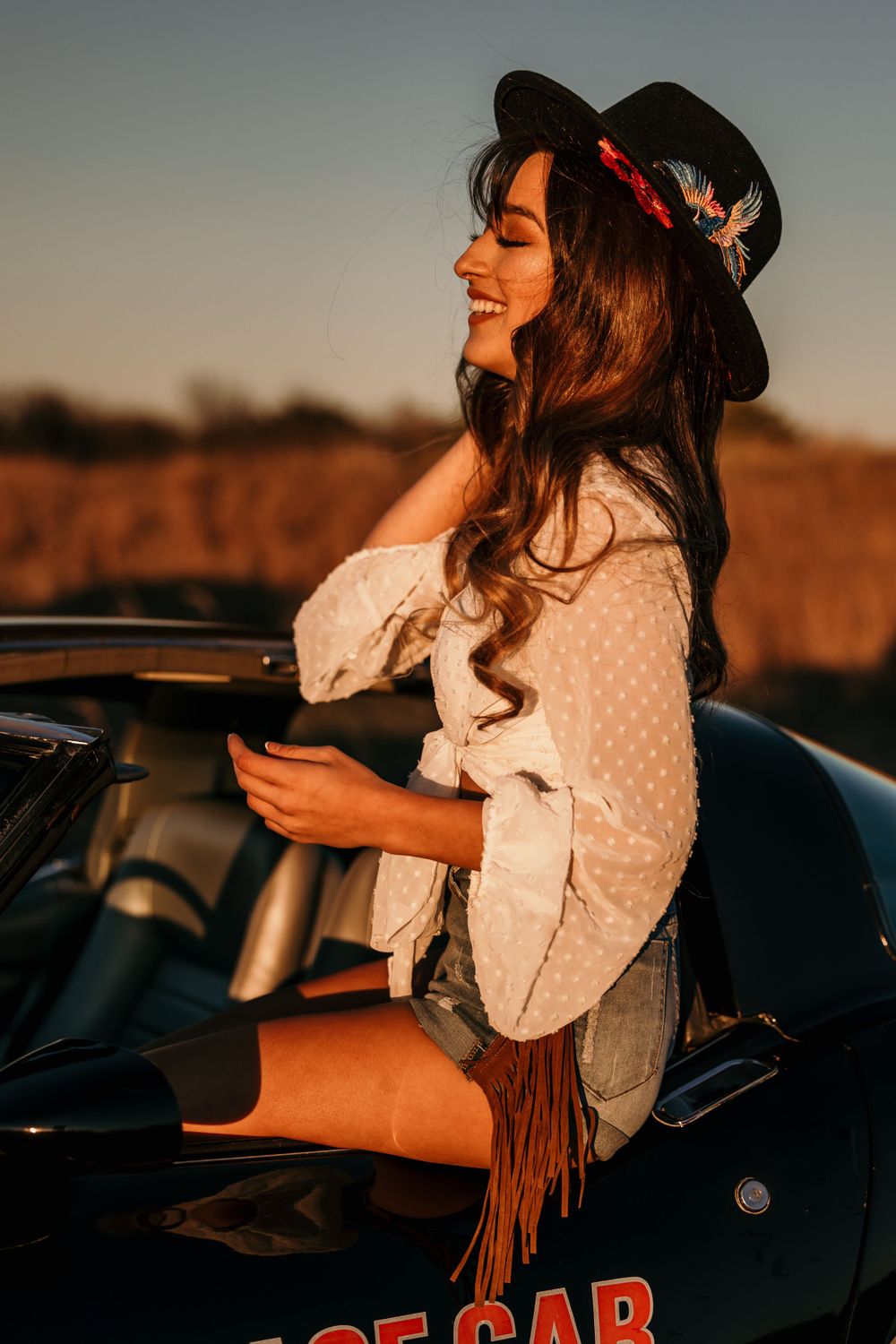 Woman in a black hat and white blouse sitting on a vintage car during golden hour.