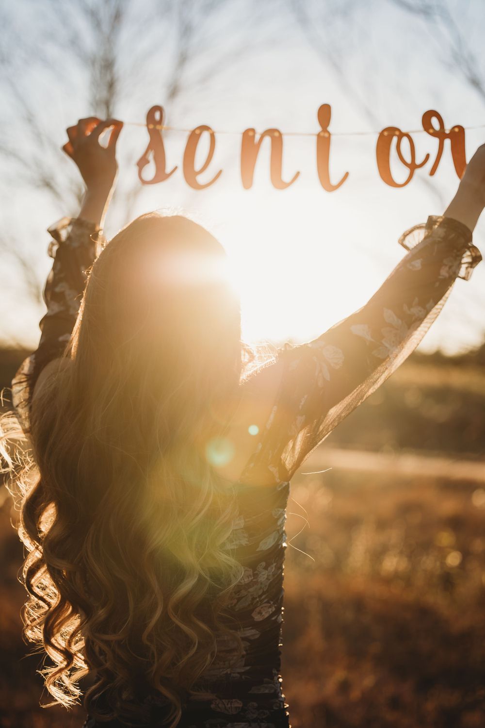 Silhouette of a person with curly hair raising their arms against sunset with 'senior' text overlay.