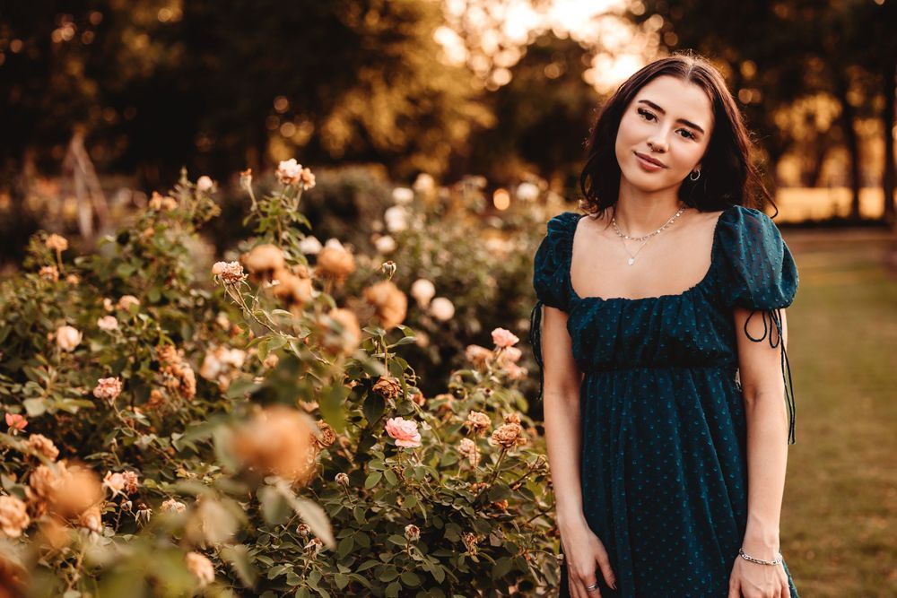 Woman in dark dress poses by blooming roses in garden at sunset.