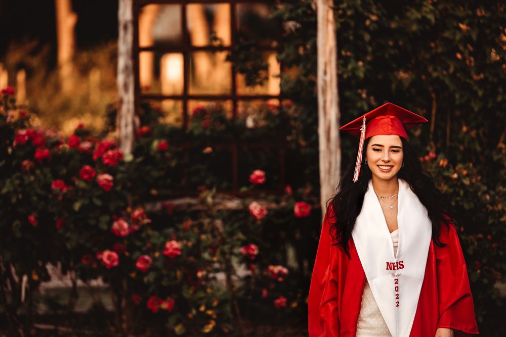Graduate in red cap and gown stands by rose garden at sunset.