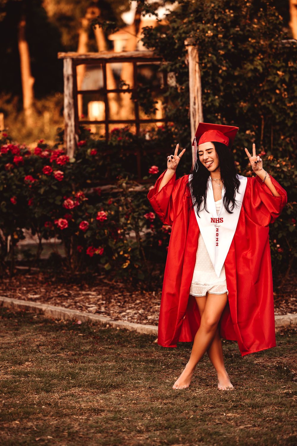 Graduate making peace signs wearing red graduation gown by rose garden.