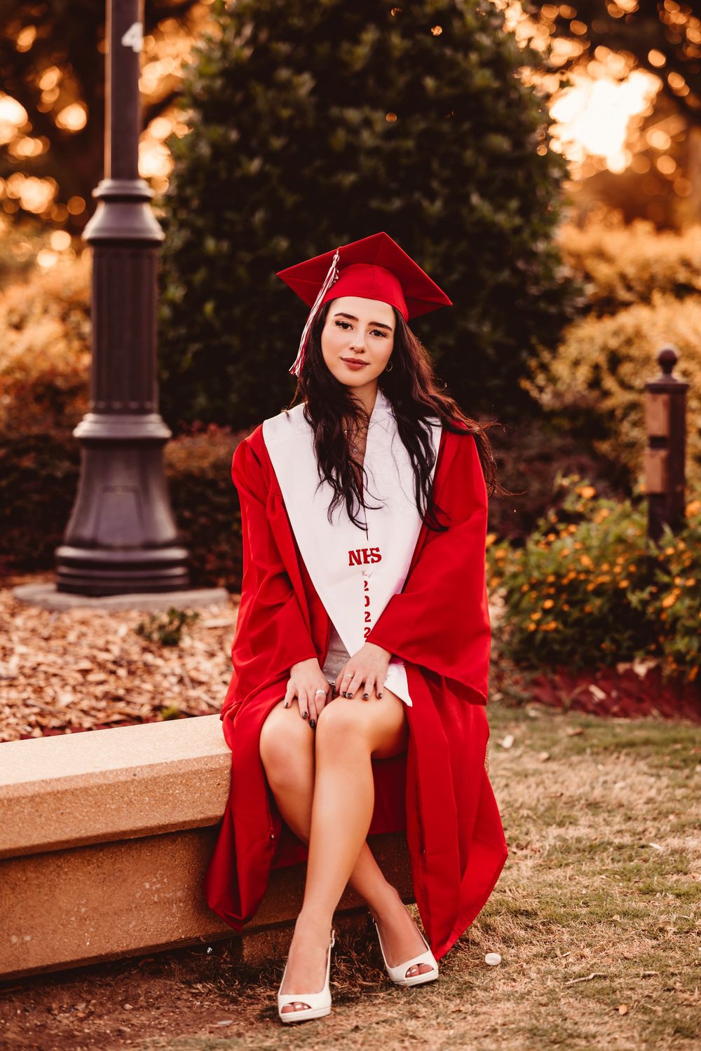 Graduate in red cap and gown sitting on stone bench at sunset.