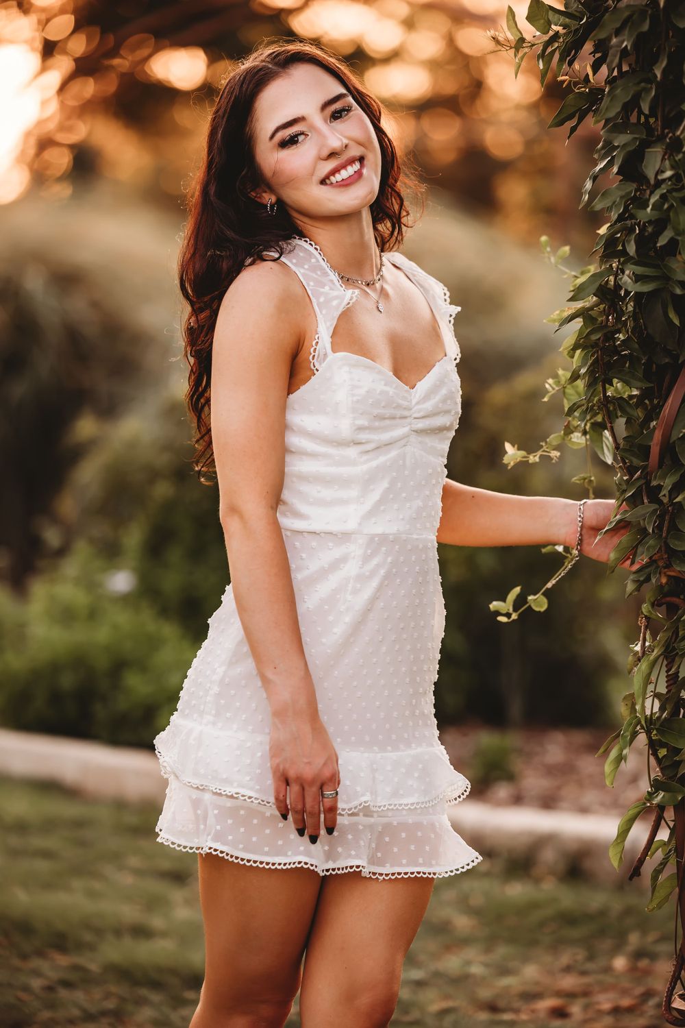 Woman in white lace dress poses outdoors with golden bokeh background.