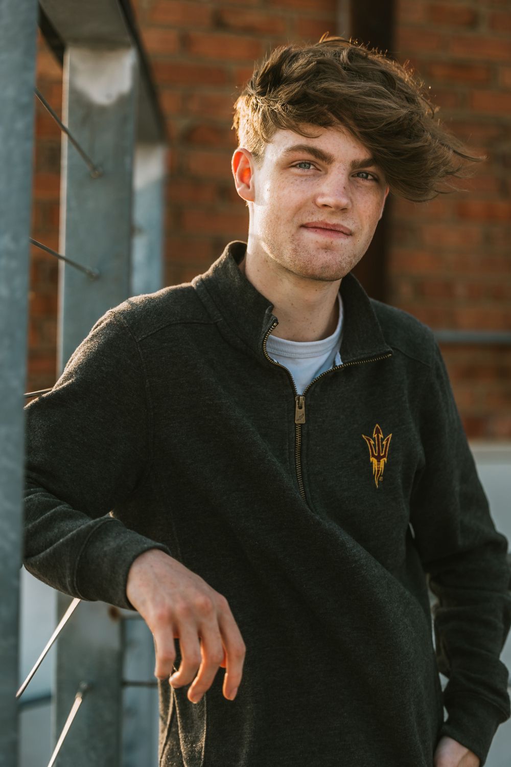 Portrait of person in black zip-up jacket with university logo against brick wall.
