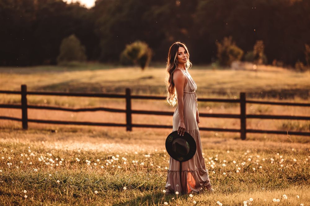 A dreamy sunset photo of a person in a flowy dress standing in a golden field with a wooden fence.