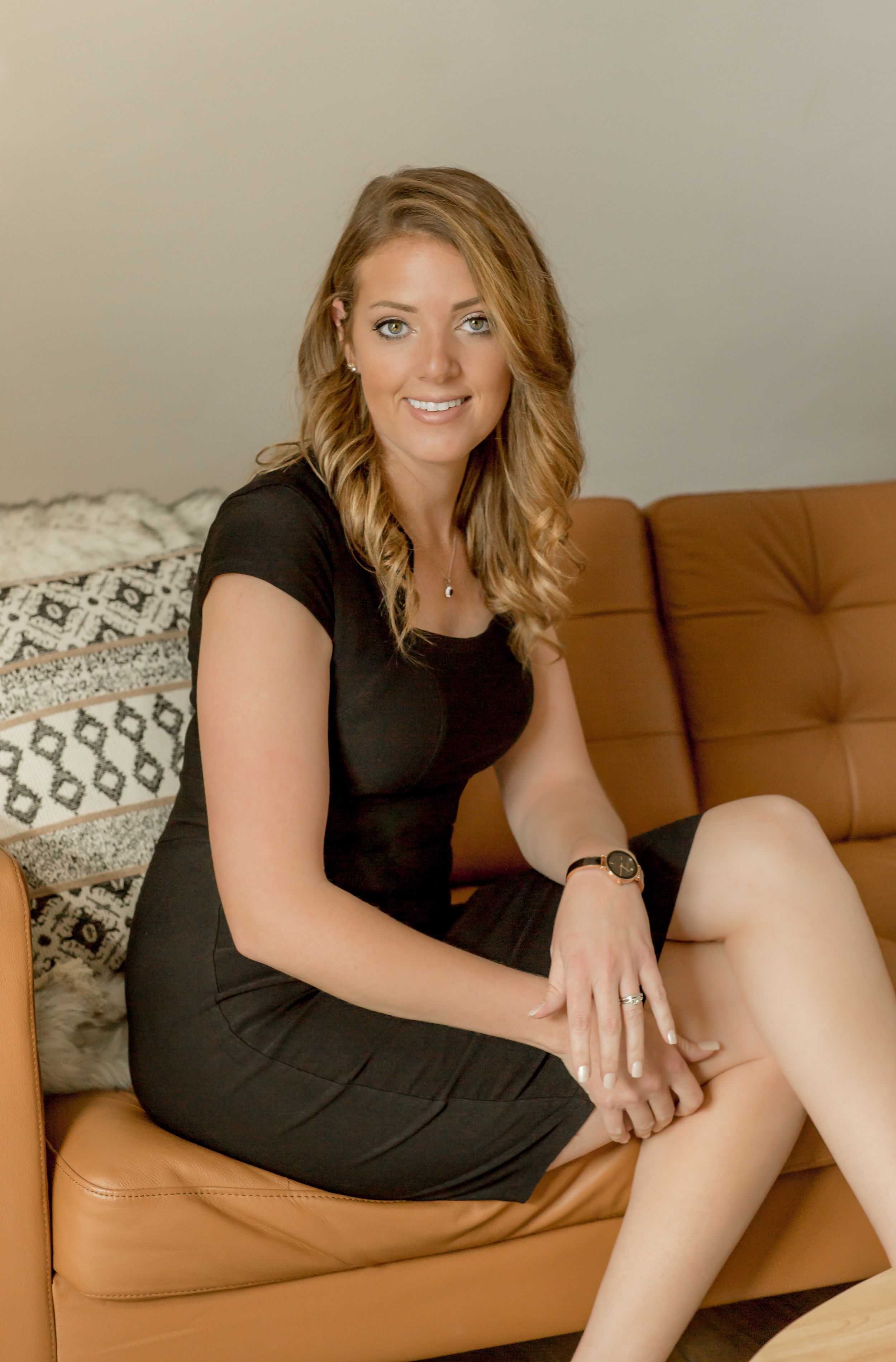 Blonde woman in black dress sitting on tan leather sofa smiling in a modern interior setting.