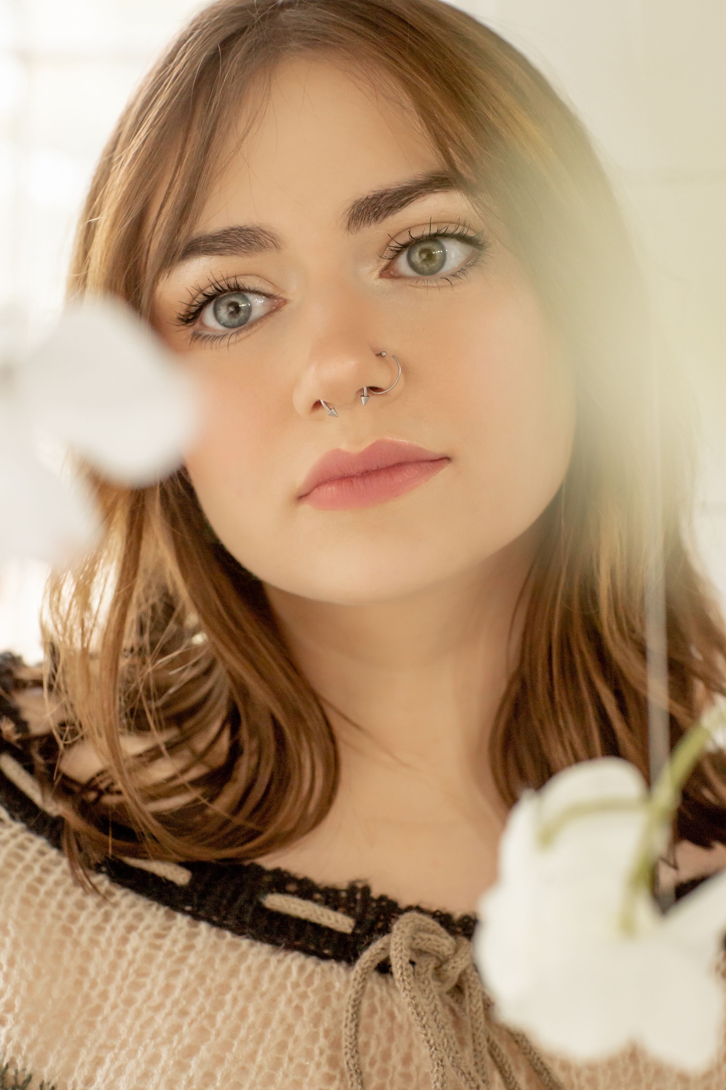 Close-up portrait of young woman with green eyes and nose ring beside white flowers in soft light.
