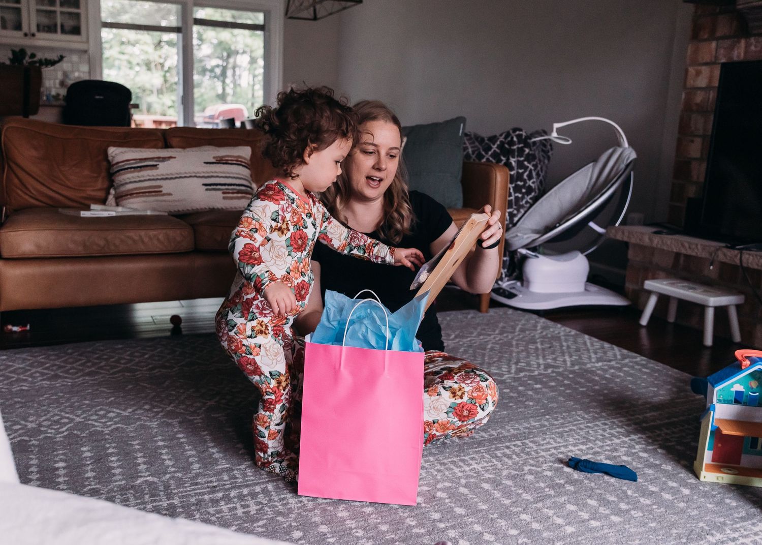 Two people explore items from a pink shopping bag while sitting on a patterned rug.