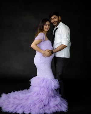 Couple in formal attire embraces in front of dark backdrop with flowing purple gown.