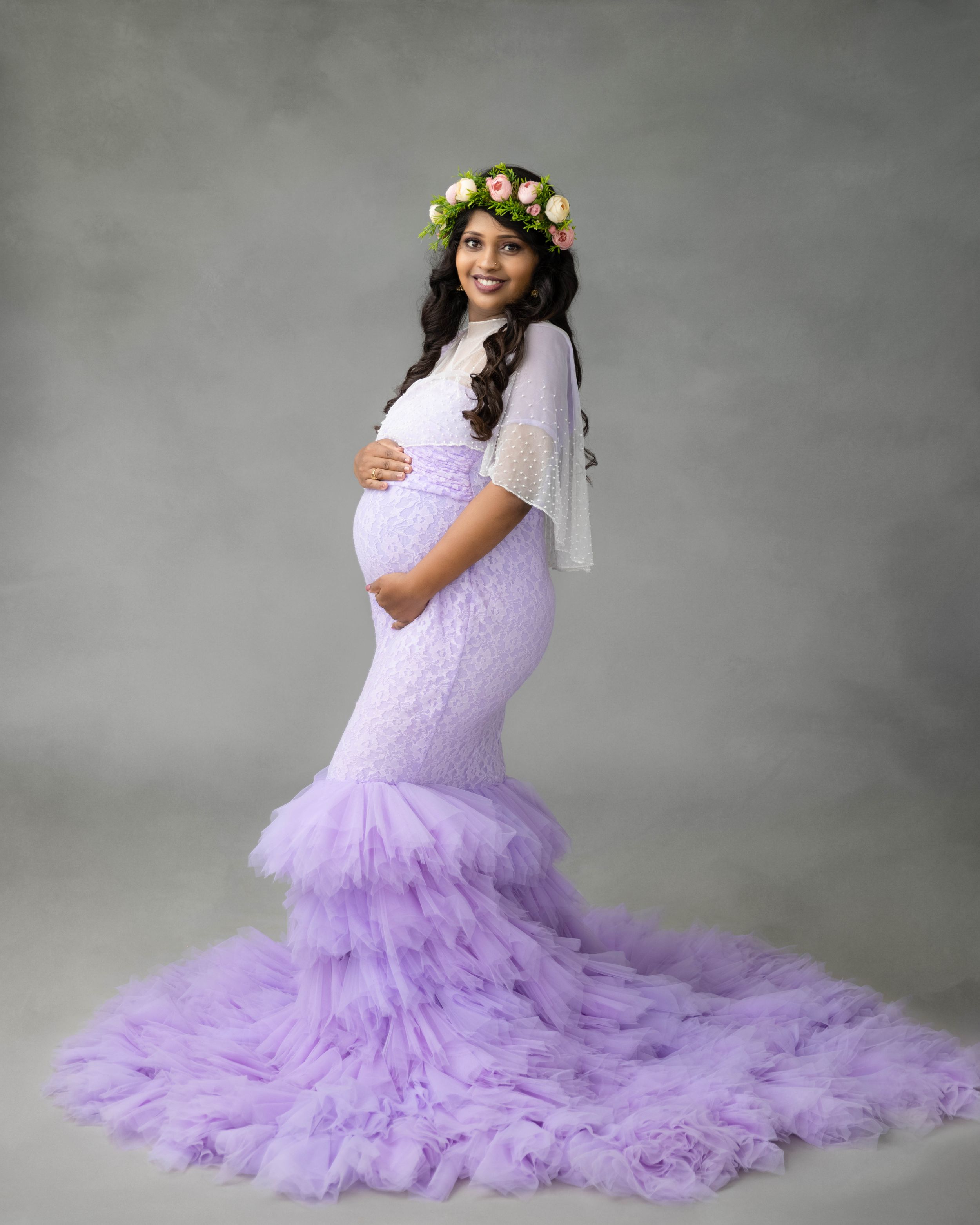 Maternity photoshoot featuring a lavender tulle mermaid gown with a floral crown against a gray backdrop.