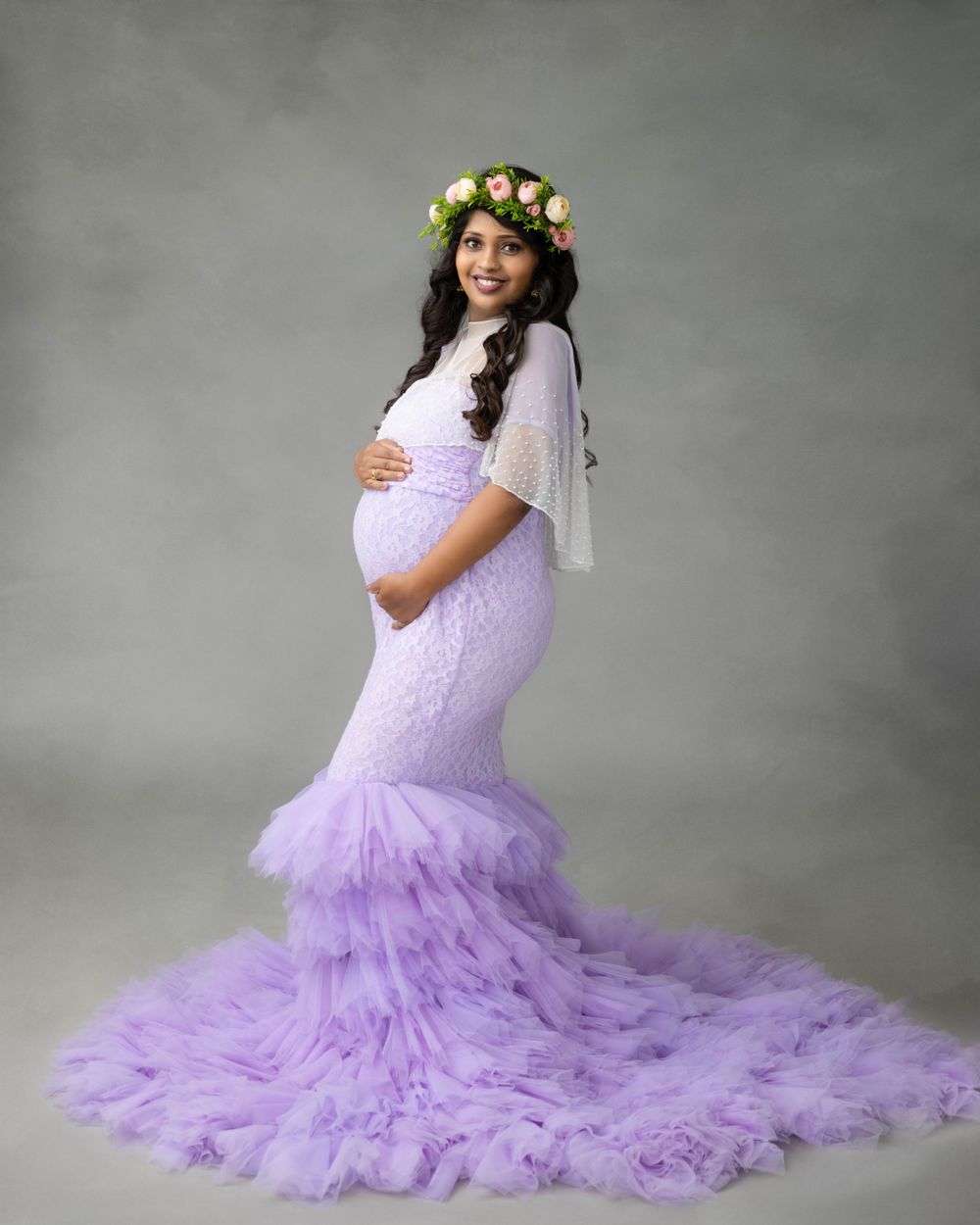 Maternity photoshoot featuring a lavender tulle mermaid gown with a floral crown against a gray backdrop.