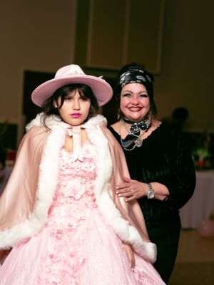 Two people posing together, one wearing a pink formal gown with fur trim and matching hat.