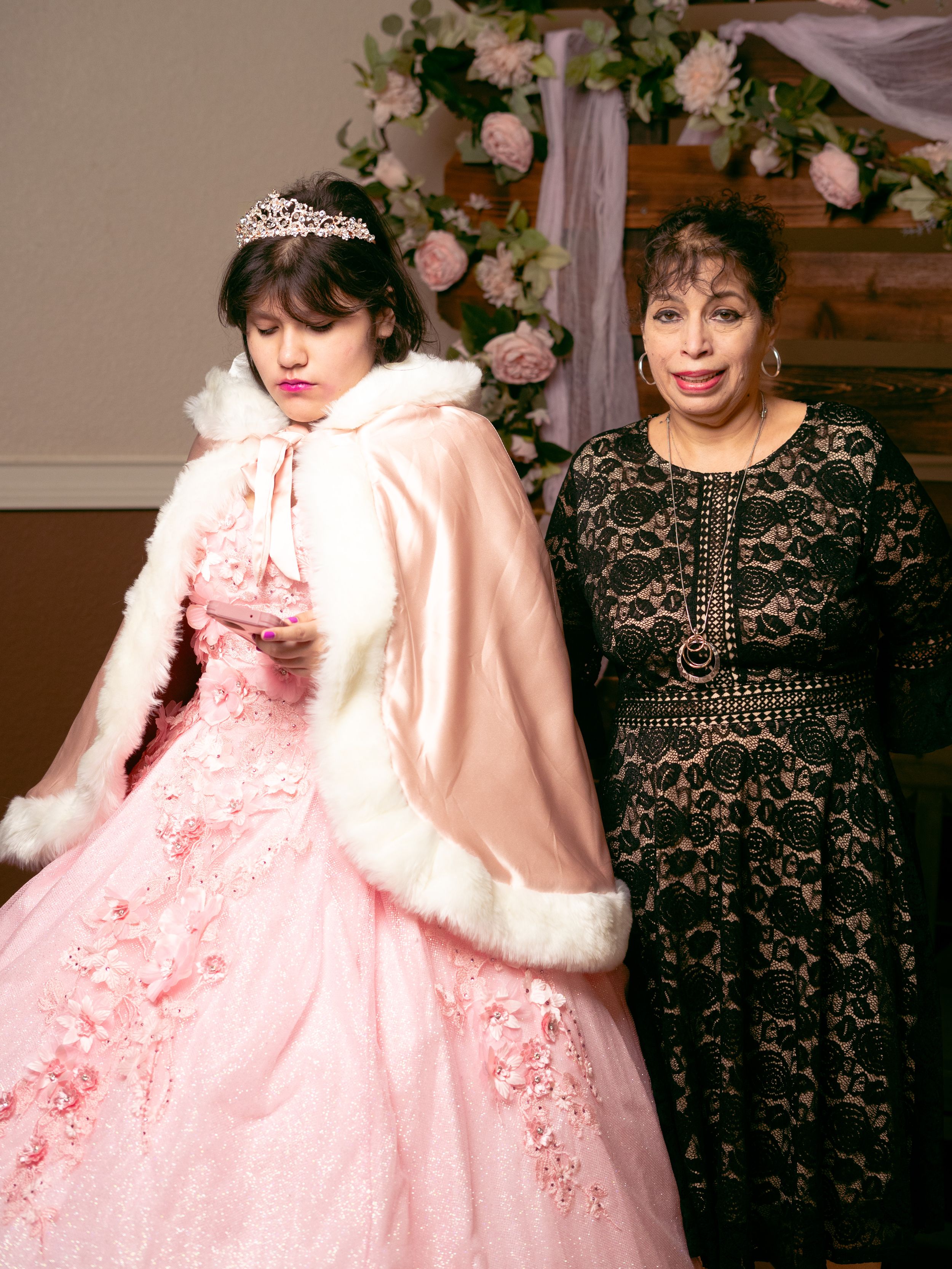 Quinceañera celebration with two guests in formal attire against a floral backdrop.