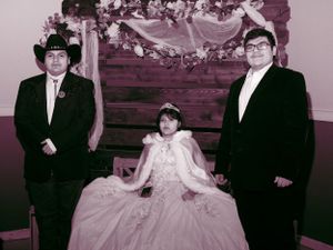 A formal group photo with two people in suits and one in a white gown against a decorated backdrop at a special event.