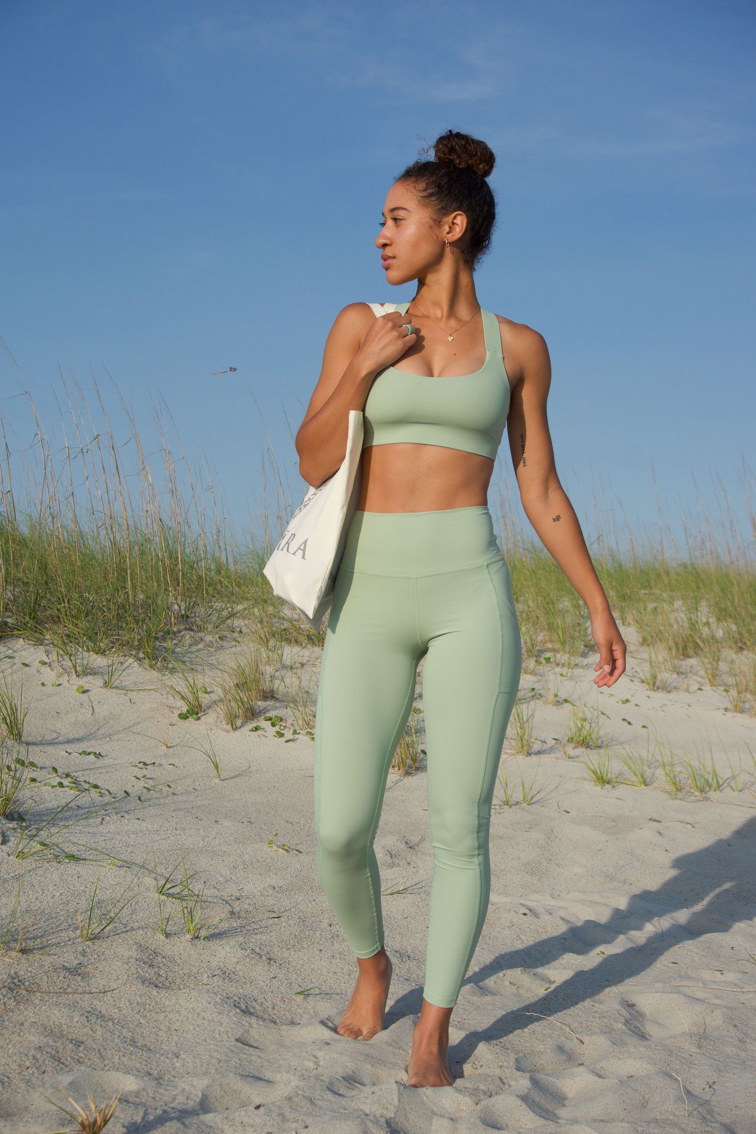 A girl walks on the beach with a canvas tote bag and light green yoga athletic wear