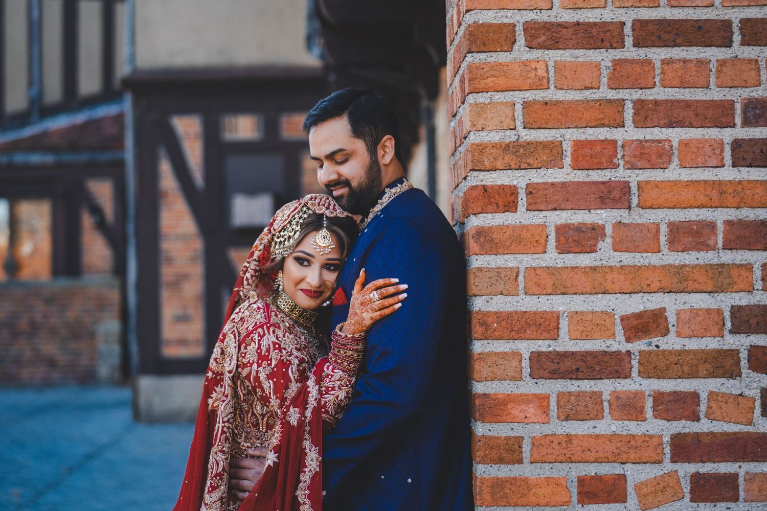 Couple in traditional South Asian wedding attire embracing against brick wall.