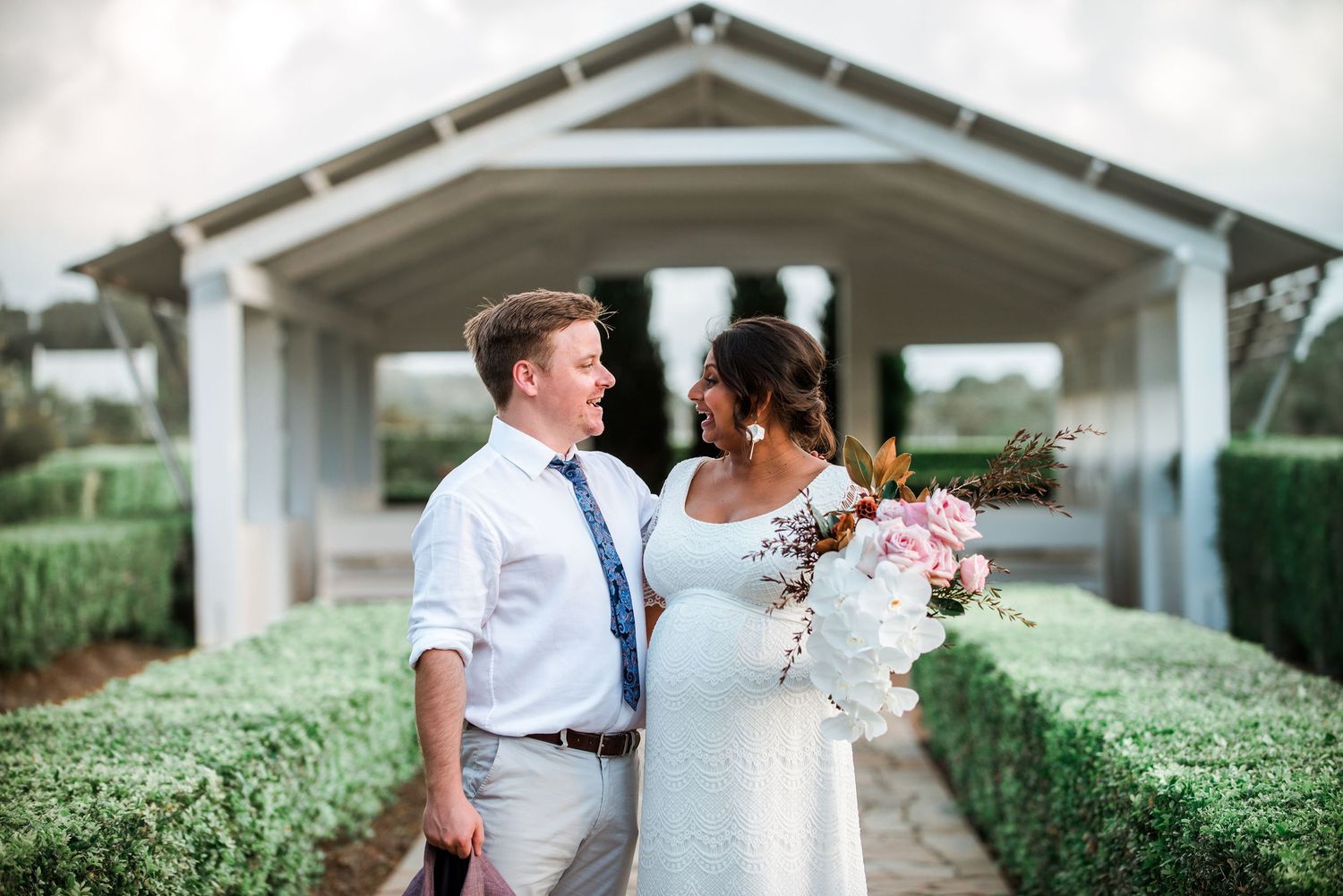 A couple in wedding attire poses in front of a white covered bridge structure.
