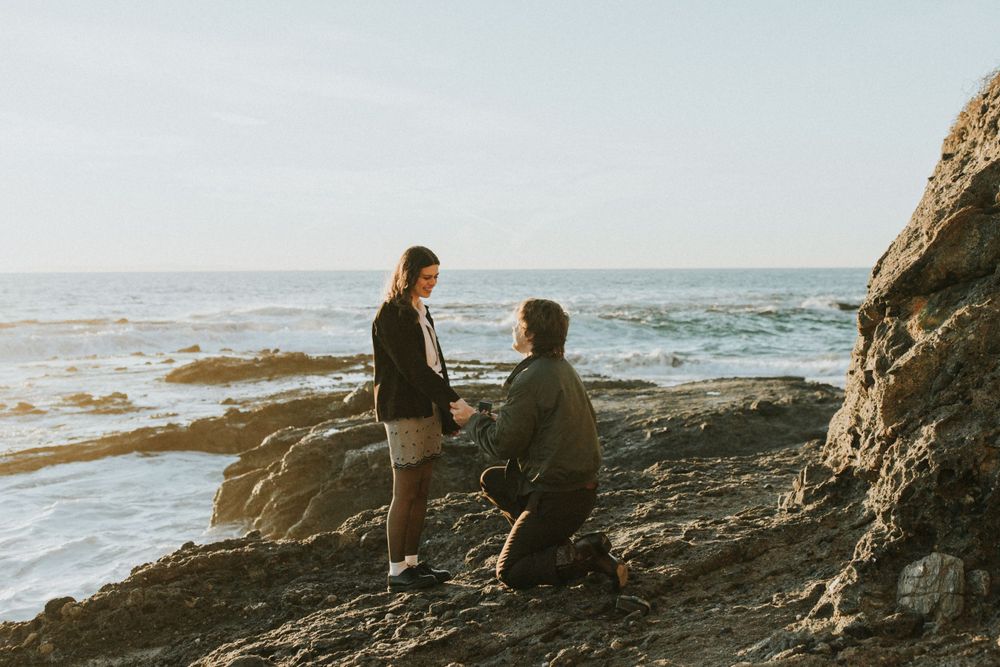A romantic proposal scene on rocky coastal cliffs with ocean waves in the background.