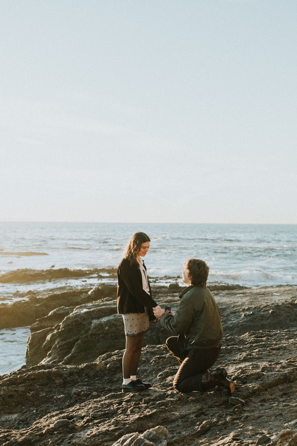 A seaside proposal moment captured on rocky cliffs during golden hour with ocean views.