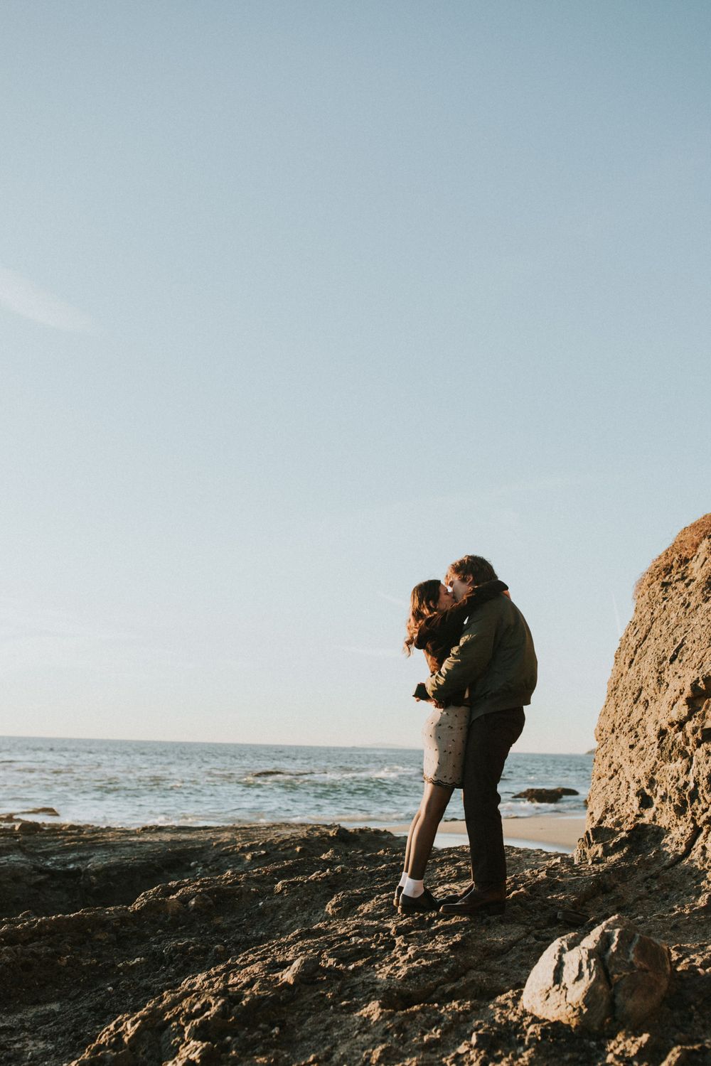 A couple embraces on coastal rocks with the ocean and blue sky in the background.