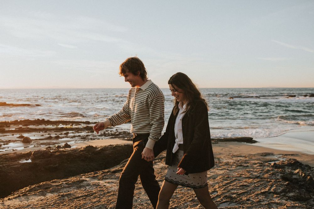 A couple walking along rocky coastal terrain during sunset with ocean waves behind them.