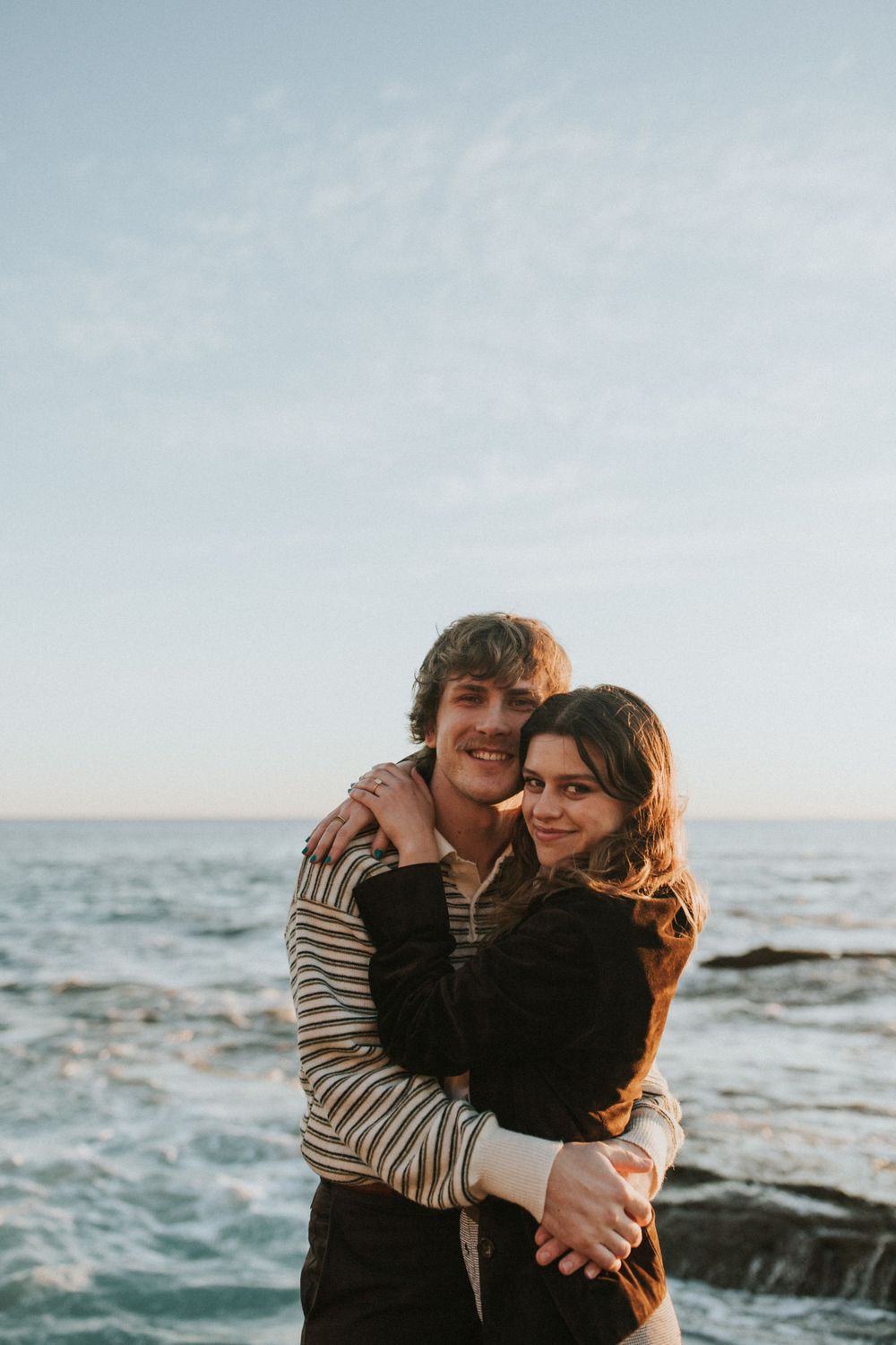 A romantic couple embraces on rocky coastal shores with ocean waves in the background during golden hour.