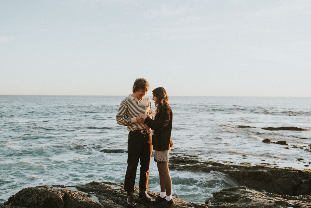 A couple stands together on coastal rocks overlooking the ocean at sunset.