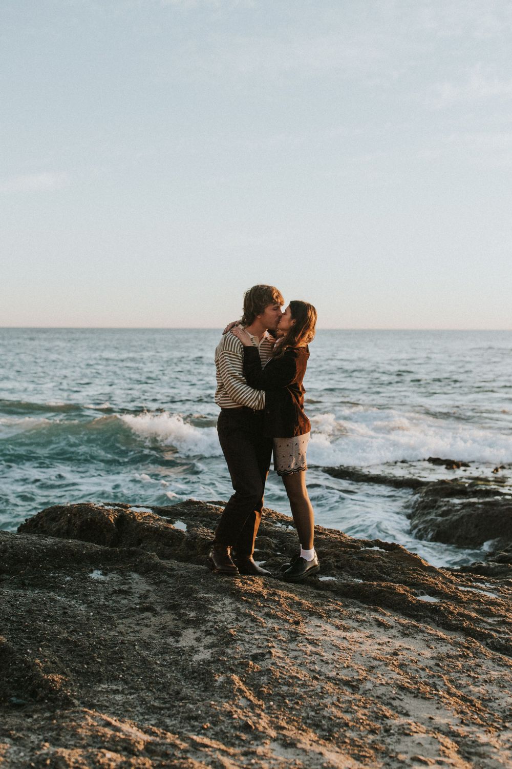 A couple shares a tender moment on rocky ocean cliffs during sunset.