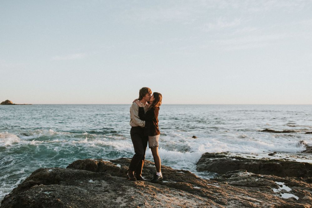 A romantic embrace on rocky coastal cliffs with crashing waves in the background.