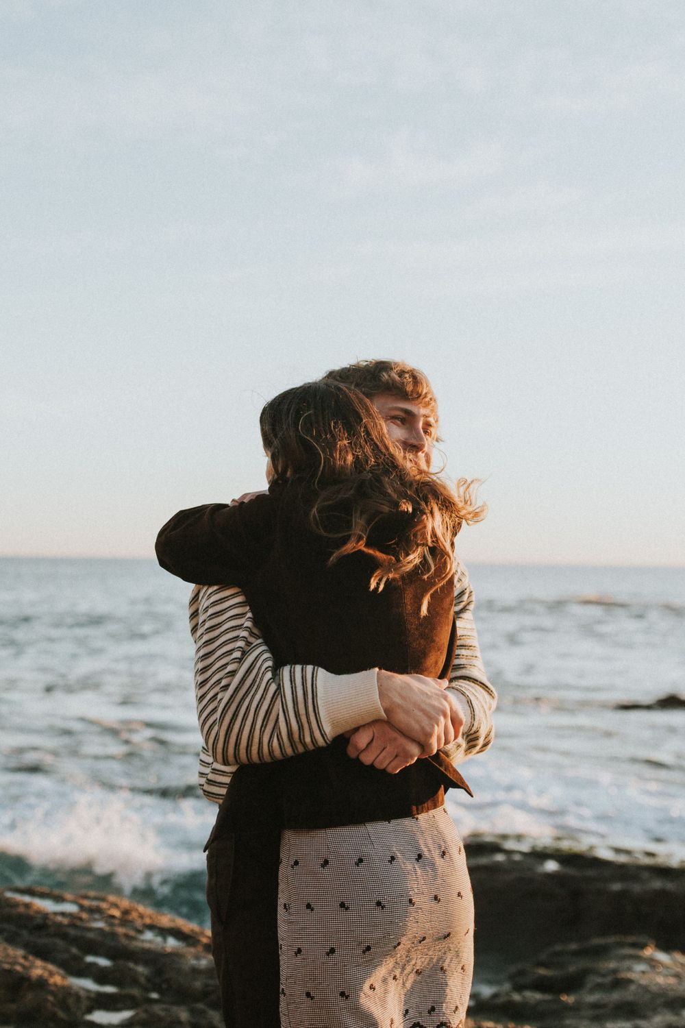 A warm embrace between two people on a rocky beach with ocean waves behind them.