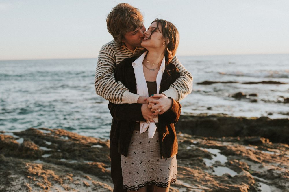 An intimate moment shared between a couple on coastal rocks with ocean waves in the background.