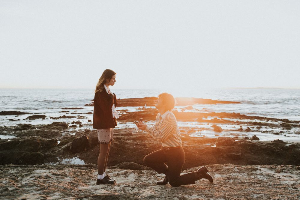A proposal scene unfolds on rocky beach during golden hour sunset.