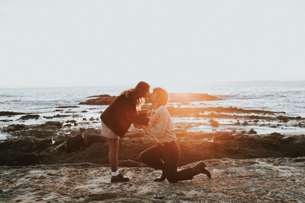 A romantic proposal moment captured at sunset on a rocky beach overlook.