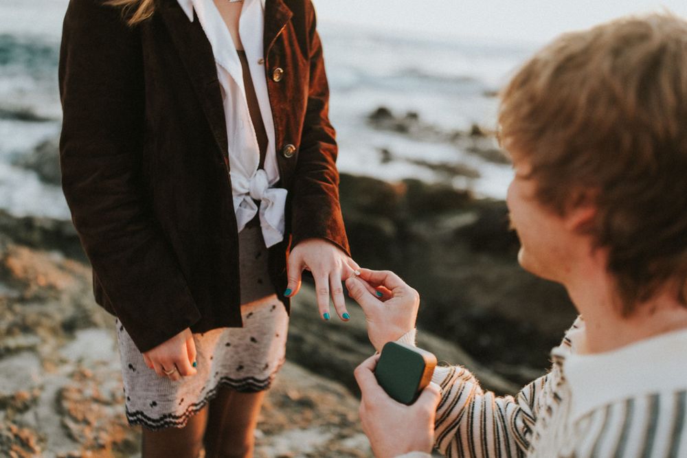 A beachside proposal scene with one person kneeling and offering a ring.