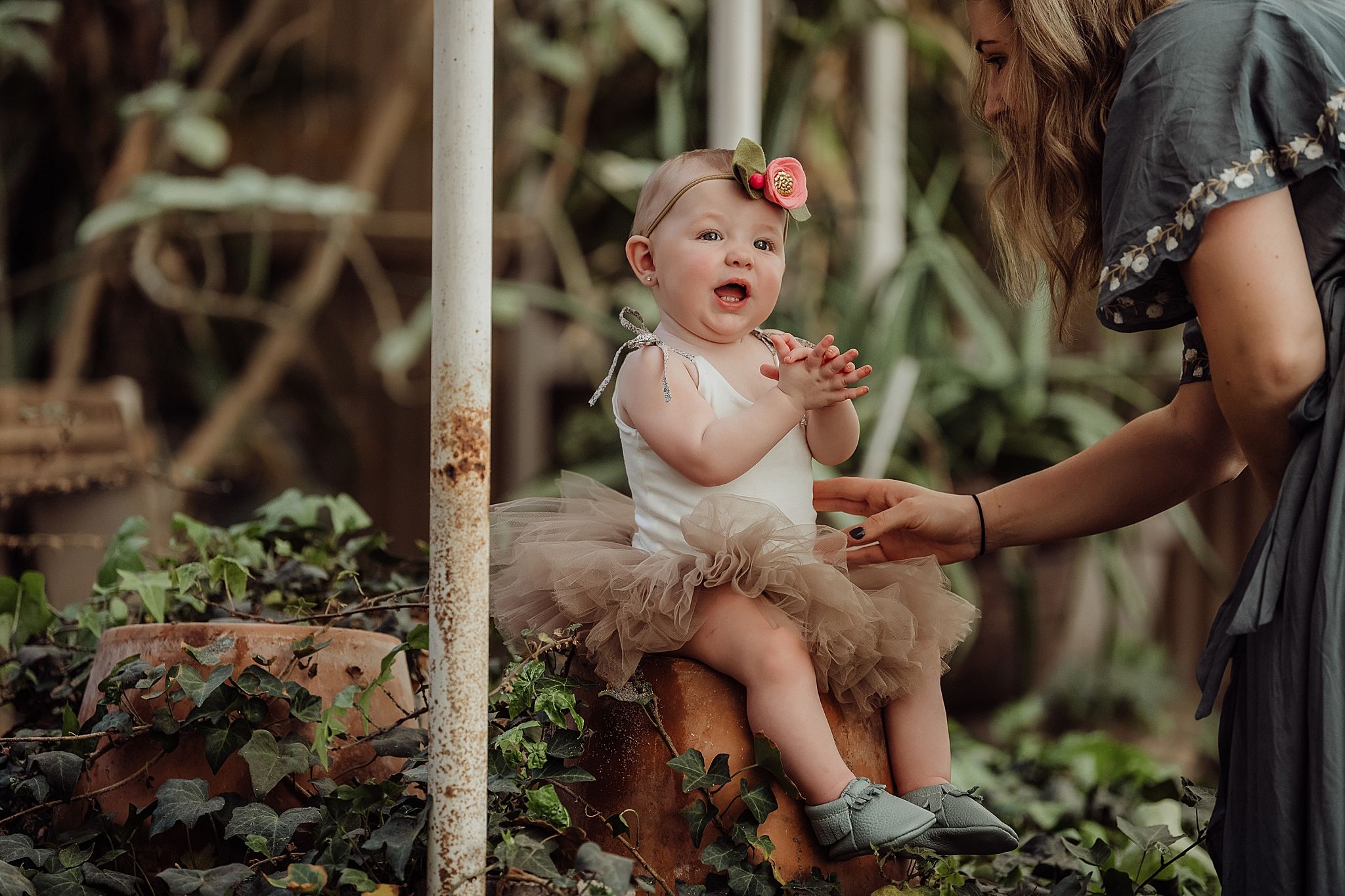 Baby in a tulle dress sits on stone steps surrounded by greenery during a garden photoshoot.
