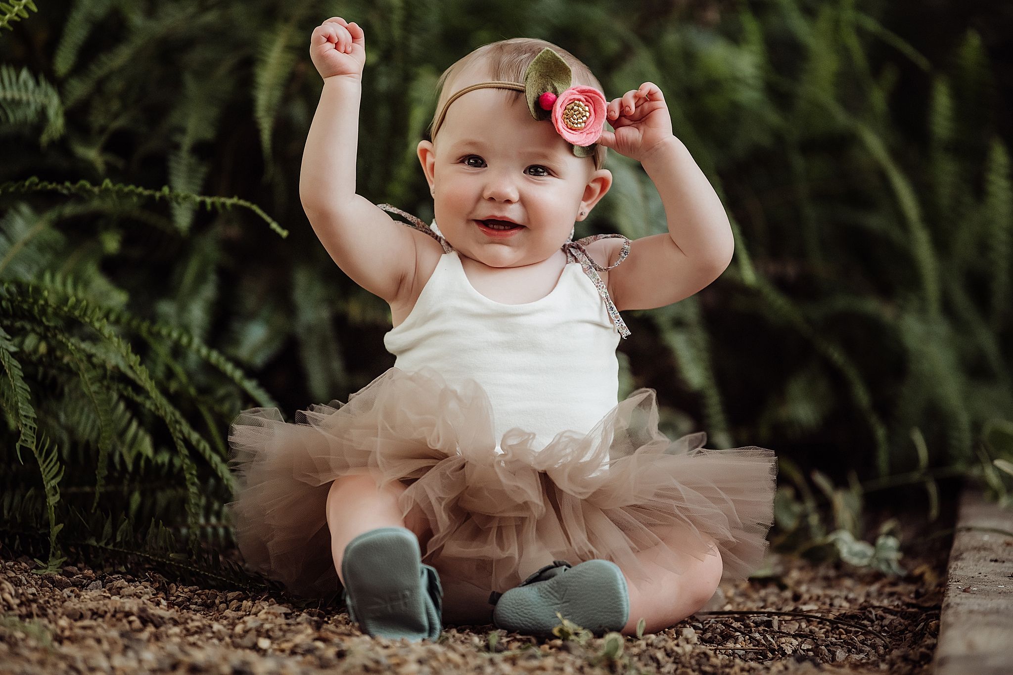 Small child wearing a tutu and floral headband sits on ground raising arms joyfully in outdoor garden setting.