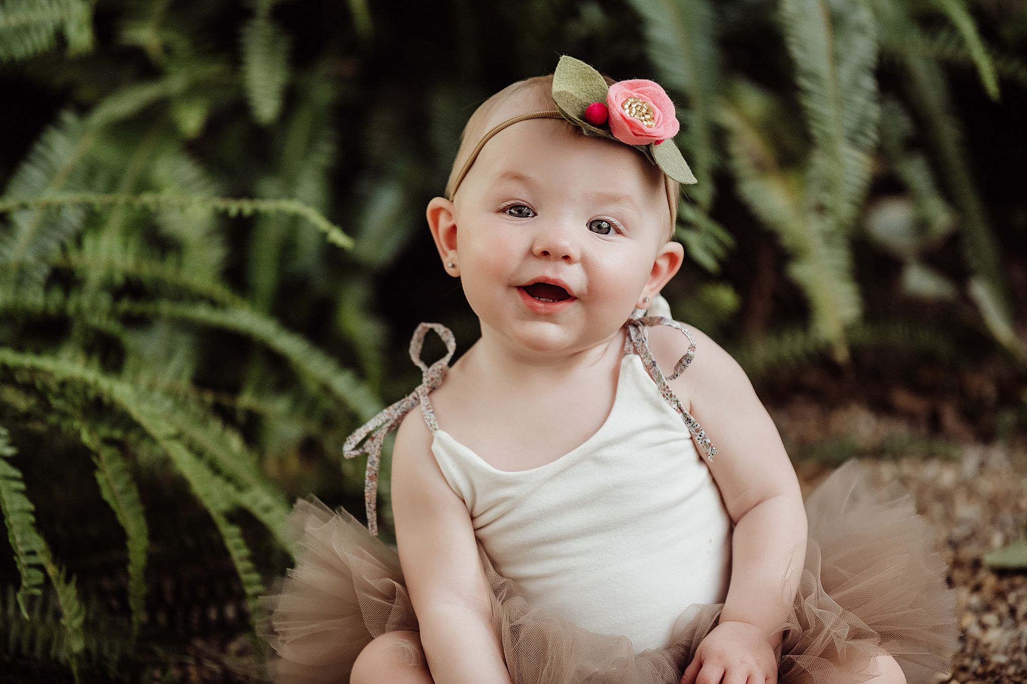 Smiling baby in cream outfit and floral headband poses against fern background.