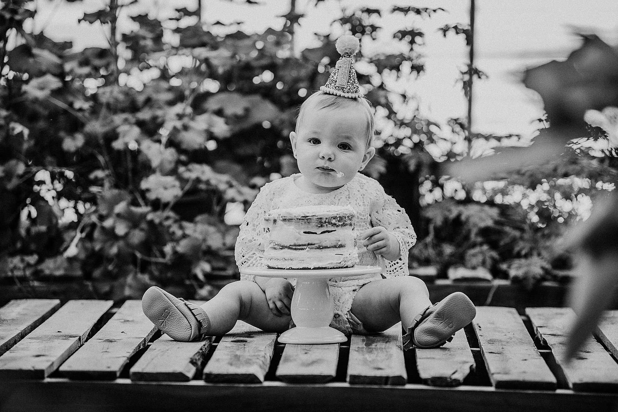 Small baby sitting with birthday cake on wooden deck in greenhouse.