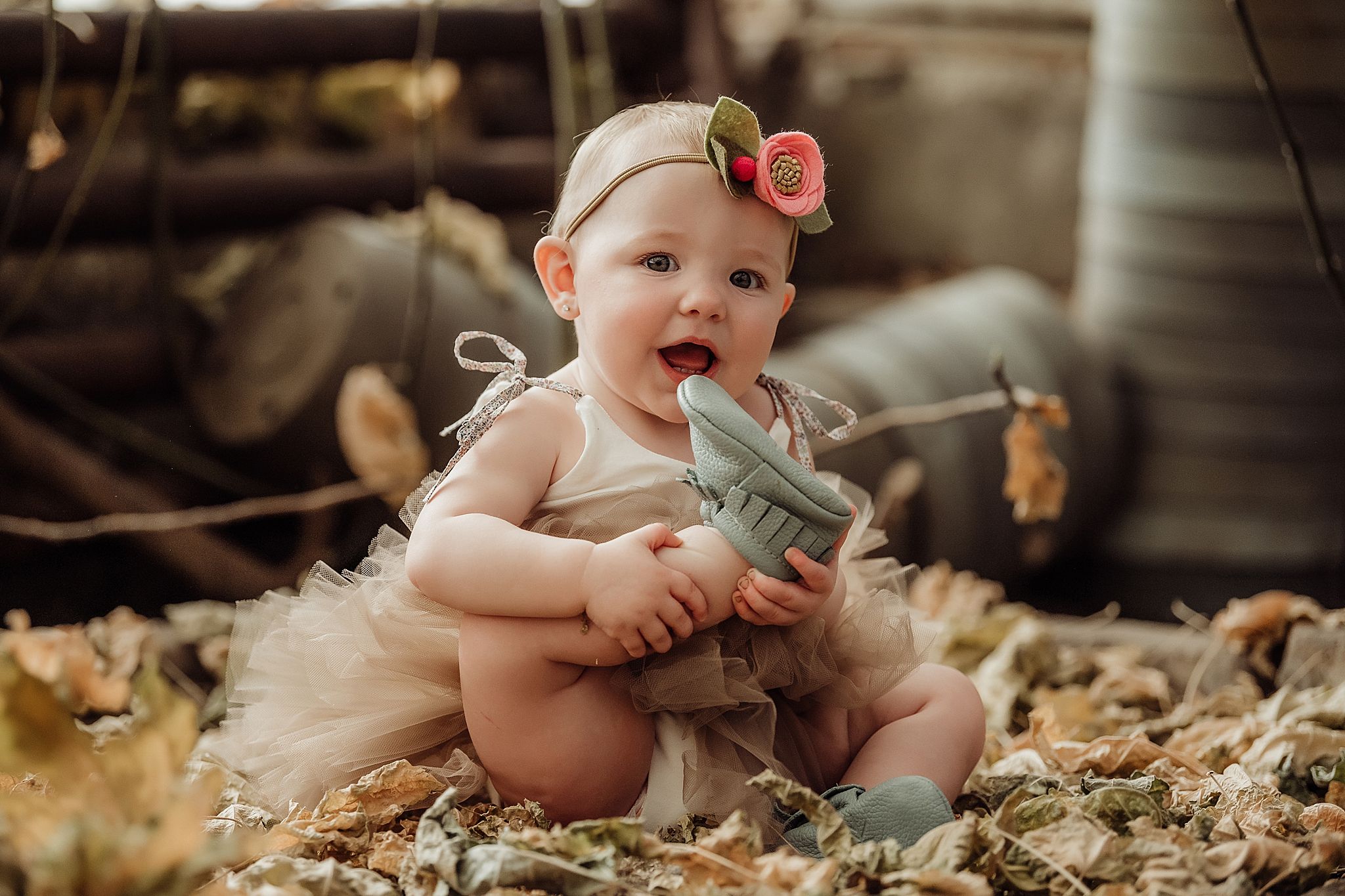 Baby in tulle dress and floral headband plays with vintage camera on autumn leaves.